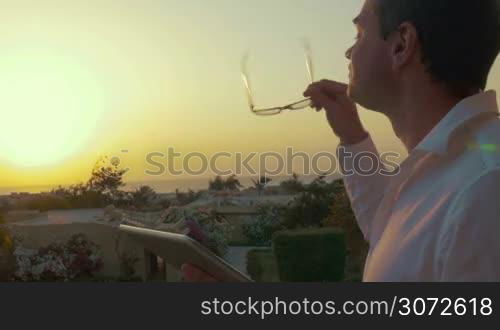 Young man is putting glasses on and writing letters in tablet PC, beautiful sunset is on the background.