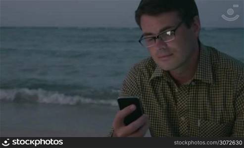 Young man is sitting by the sea and texting message in smartphone in the evening.