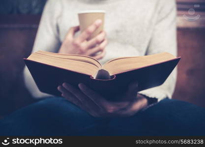 Young man is sitting on a sofa and reading a book while holding a paper cup