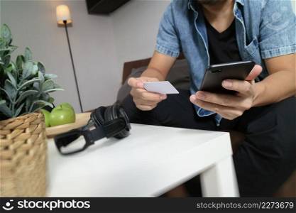 young man is sitting on a sofa shopping on line with smart phone is listening music with headphones is holding a credit card on his hand at home,omni channel concept