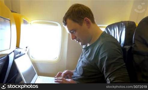 Young man is sitting on the plane with laptop on his knees. He&acute;s typing something, clock-watching and looking out of the window.