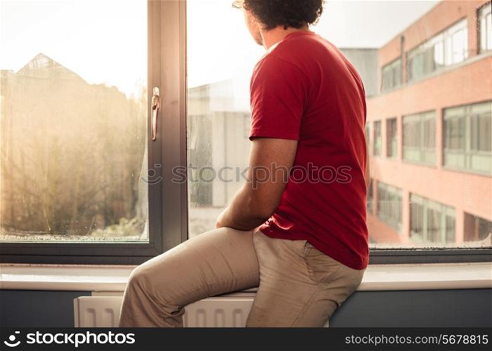 Young man is sitting on window silll and looking out at sunset