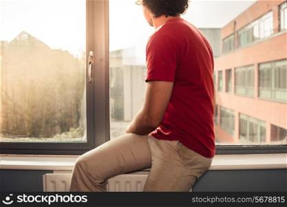 Young man is sitting on window silll and looking out at sunset