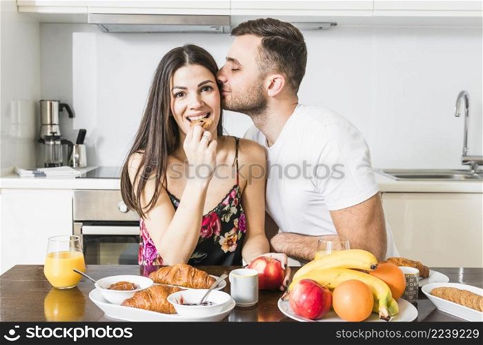 young man kissing her girlfriend eating cookies with fruits croissant table kitchen