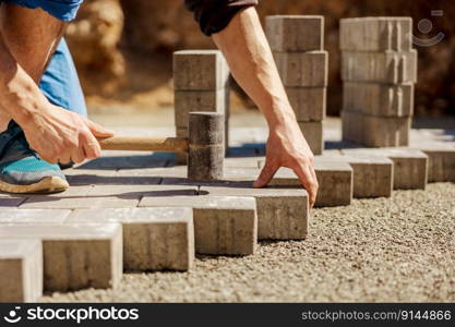 Young man laying gray concrete paving slabs in house courtyard on gravel foundation base. Master lays paving stones. Garden brick pathway paving by professional paver worker. Repairing sidewalk. Young man laying gray concrete paving slabs in house courtyard on gravel foundation base. Master lays paving stones. Garden brick pathway paving by professional paver worker. Repairing sidewalk.