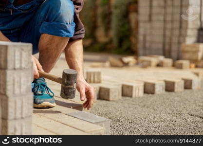 Young man laying gray concrete paving slabs in house courtyard on gravel foundation base. Master lays paving stones. Garden brick pathway paving by professional paver worker. Repairing sidewalk. Young man laying gray concrete paving slabs in house courtyard on gravel foundation base. Master lays paving stones. Garden brick pathway paving by professional paver worker. Repairing sidewalk.