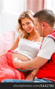 Young man listening baby in his pregnant wifes tummy using stethoscope&#xA;