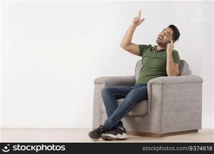 Young man listening music on headphones and pointing above with his index finger while sitting on sofa