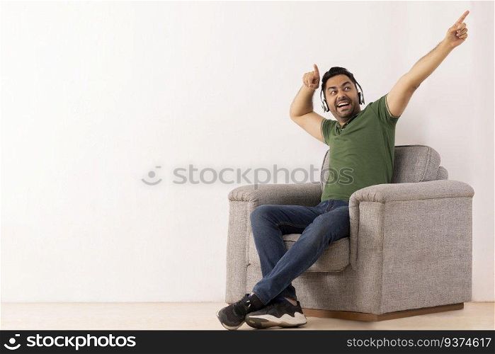Young man listening music on headphones and pointing away with his index fingers while sitting on sofa