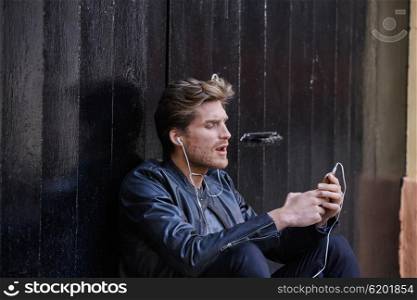 Young man listening music with smartphone earphones sitting in the street black door