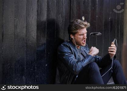 Young man listening music with smartphone earphones sitting in the street black door