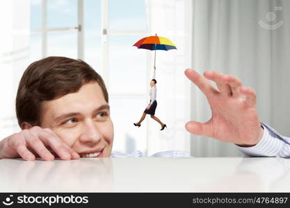 Young man looking from under table on businesswoman flying on umbrella. He is big boss