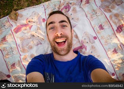 Young man lying on a sheet in the park and taking selfie picture with goofy smile.