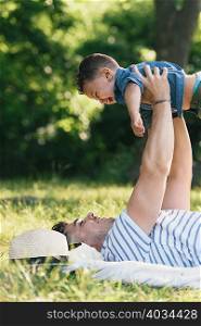 Young man lying on back holding up toddler son in Pelham Bay Park, Bronx, New York, USA
