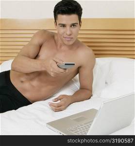 Young man lying on the bed using a remote control with a laptop in front of him