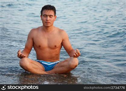 Young man meditating on beach, sitting on edge of water.