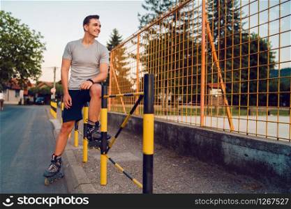 Young man on roller blades skates sitting on the fence wall looking waiting in a summer day evening rollerblade