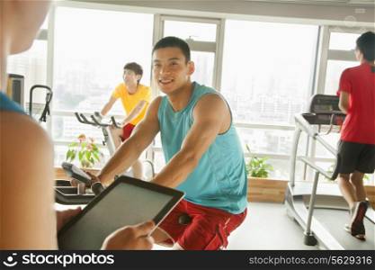 Young man on stationary bike exercising with his personal trainer