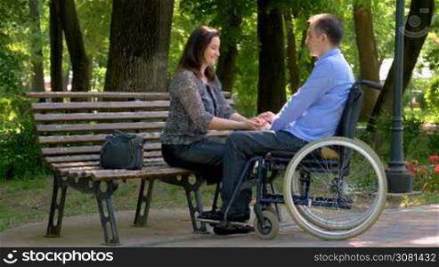 Young man on wheelchair talking with his wife in park. Wife encouraging his disabled husband. Slow motion