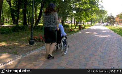 Young man on wheelchair walking with his wife outdoors in park.