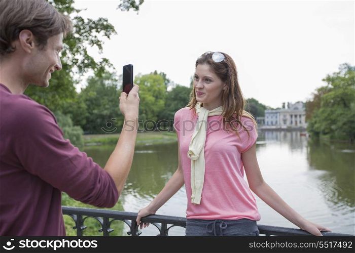 Young man photographing woman through cell phone at lakeside