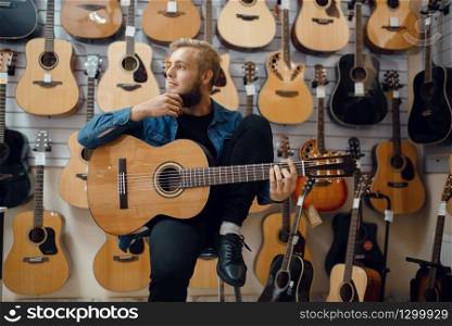 Young man plays on acoustic guitar in music store. Assortment in musical instruments shop, male musician buying equipment. Young man plays on acoustic guitar in music store