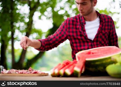 young man putting spices on raw meat for barbecue grill during outdoor french dinner party near the river on beautiful summer evening in nature