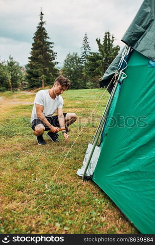 Young man putting up a tent on c&ing during summer vacation trip. Teenager putting the stakes into grassy ground using hammer. Young man putting up a tent on c&ing during summer vacation trip