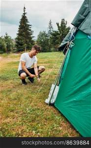 Young man putting up a tent on c&ing during summer vacation trip. Teenager putting the stakes into grassy ground using hammer. Young man putting up a tent on c&ing during summer vacation trip