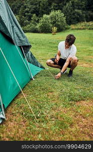 Young man putting up a tent on c&ing during summer vacation trip. Teenager putting the stakes into grassy ground using hammer. Young man putting up a tent on c&ing during summer vacation trip