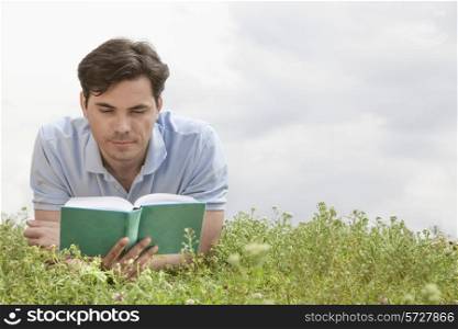 Young man reading book while lying on grass against sky