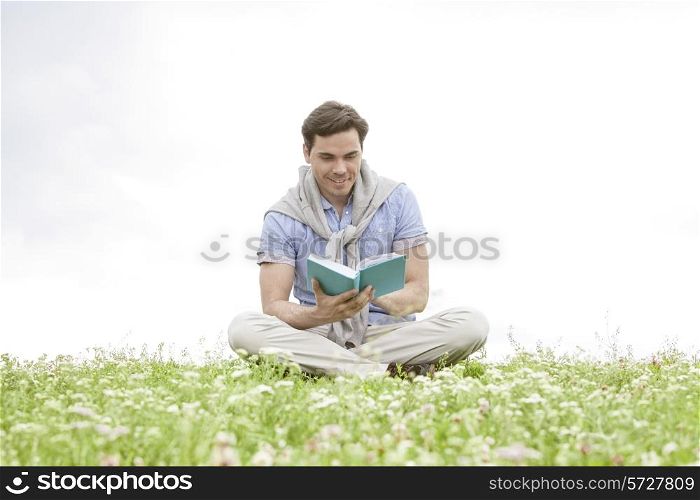Young man reading book while sitting on grass against sky