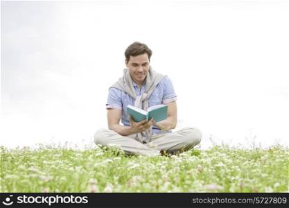 Young man reading book while sitting on grass against sky
