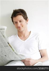 Young man reading newspaper in bed and smiling