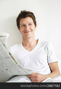 Young man reading newspaper in bed looking to side and smiling