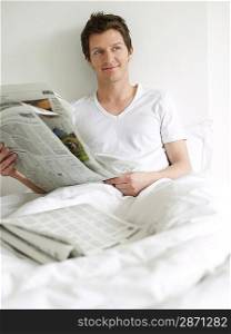 Young man reading newspaper in bed looking to side and smiling