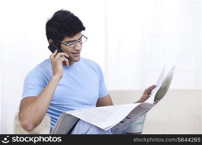 Young man reading newspaper while talking on mobile phone