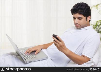 Young man reading text message on cell phone while working on laptop