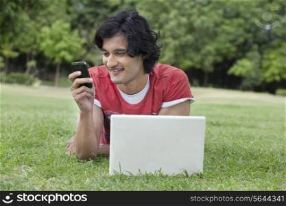 Young man reading text message with laptop in front lying on grass