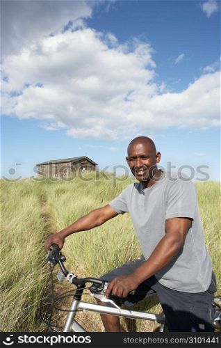 Young Man Riding Mountain Bike By Dunes With Old Beach Hut In Distance