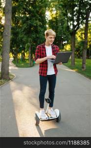 Young man riding on mini gyro board with laptop, summer park. Outdoor recreation with electric gyroboard. Eco transport with balance technology, electrical gyroscope vehicle