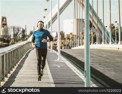 young man running on a bridge in a blue shirt and listening to music with a pair of headphones.