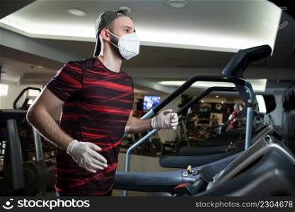 Young man running on treadmill in indoor gym,wearing protective face mask   rubber latex gloves,COVID-19 pandemic prevention of spread   transfer of Coronavirus,staying fit in lockdown concept in US