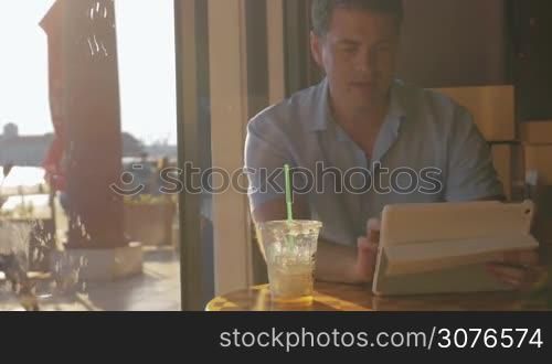 Young man searching something in white tablet computer drinking iced tea from tubule in clear plastic glass in cafe with showing window at summer sun day.