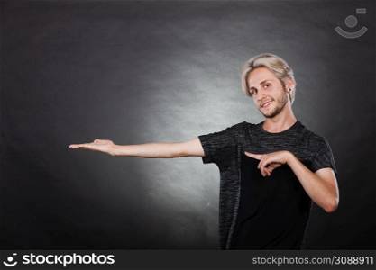 Young man showing presenting. Stylish guy pointion with finger, holding empty hand palm copyspace for product. Fashion, advertising concept. Studio shot on dark. Guy holding empty hand copy space for product