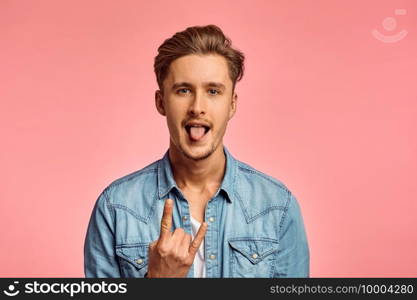 Young man shows his tongue, pink background, emotion. Face expression, male person looking on camera in studio, emotional concept, feelings. Man shows his tongue, pink background, emotion