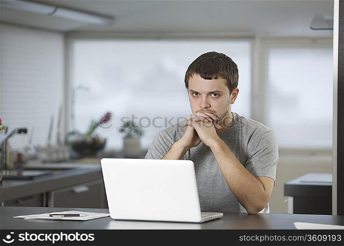 Young man sits leaning on elbows at laptop in kitchen