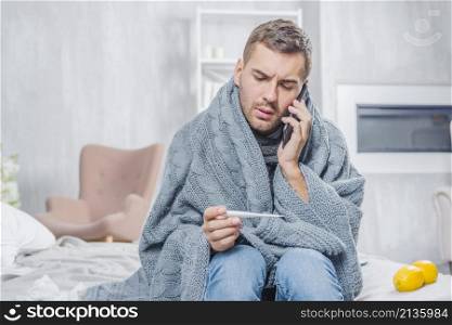 young man sitting bed wrapped scarf looking thermometer talking mobile phone