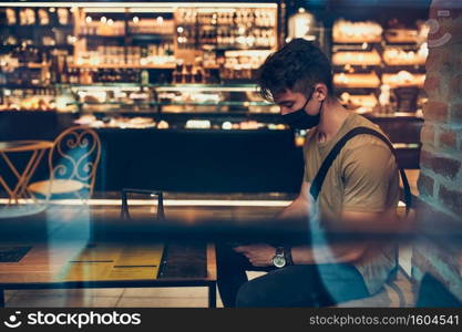 Young man sitting in coffee shop at store front in the city center in the evening, wearing the face mask to avoid virus infection and to prevent the spread of disease in time of coronavirus