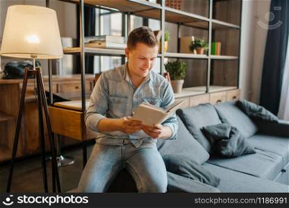 Young man sitting in couch and reading a book. Male person reads in living room, floor lamp and shelf on background